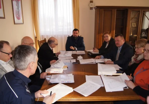 Grupa urzędników siedzi przy dużym drewnianym stole, omawiając dokumenty podczas posiedzenia Zarządu Powiatu Płockiego.