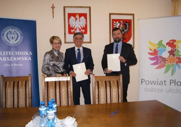 Trzy osoby w garniturach stoją za stołem, trzymając podpisane dokumenty podczas podpisywania porozumienia o współpracy między Powiatem Płockim a Politechniką Warszawską Filia w Płocku.