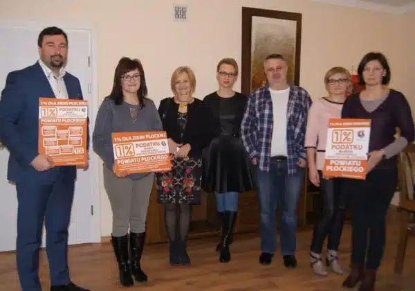 Grupa osób trzymających plakaty promujące akcję „1% Dla Ziemi Płockiej” w przytulnym wnętrzu.