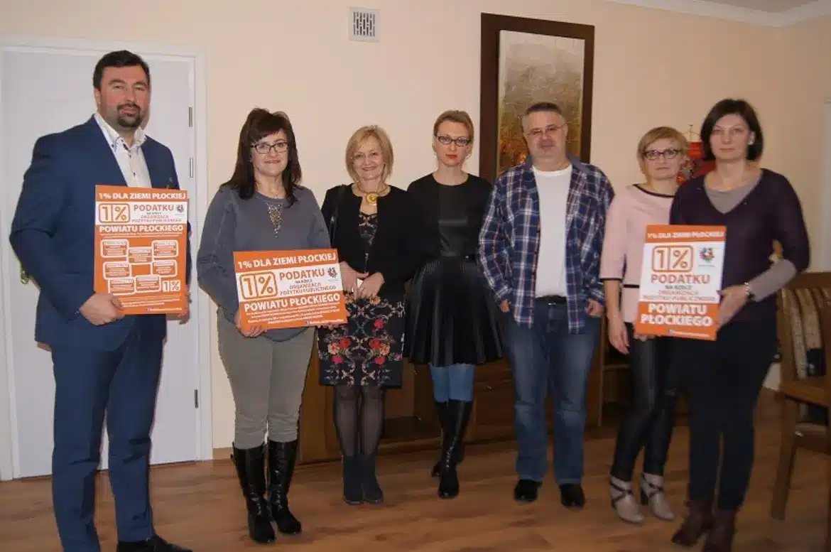 Grupa osób trzymających plakaty promujące akcję „1% Dla Ziemi Płockiej” w przytulnym wnętrzu.