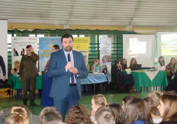 Na zdjęciu przedstawiono mężczyznę w garniturze, który przemawia do zgromadzonej grupy na IX Targach Edukacji i Pracy Szkół Ponadgimnazjalnych w Małej Wsi, z różnymi stoiskami informacyjnymi w tle.