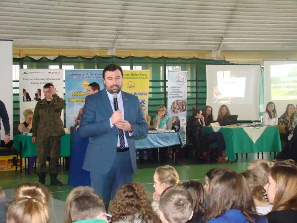 Na zdjęciu przedstawiono mężczyznę w garniturze, który przemawia do zgromadzonej grupy na IX Targach Edukacji i Pracy Szkół Ponadgimnazjalnych w Małej Wsi, z różnymi stoiskami informacyjnymi w tle.