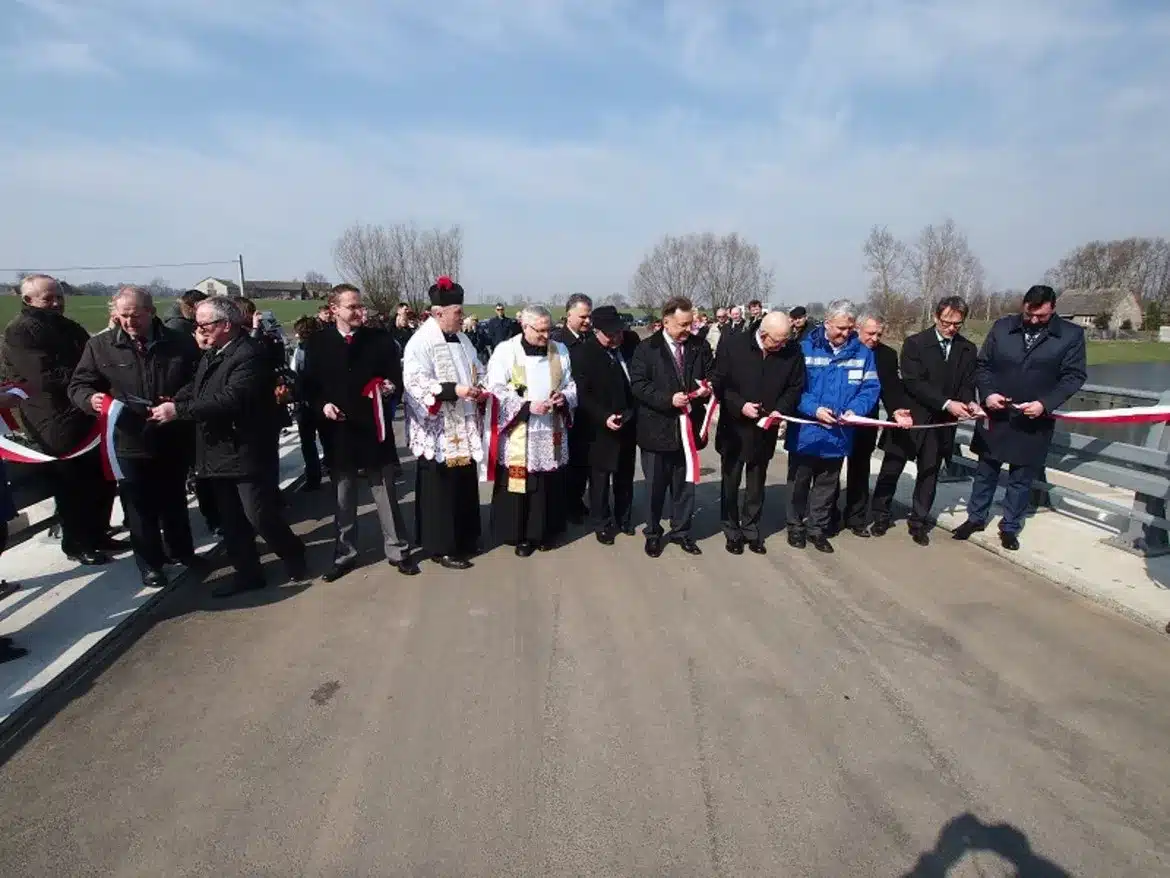 Na zdjęciu widoczna jest grupa mężczyzn, w tym duchowni i samorządowcy, trzymających wstęgę podczas ceremonii otwarcia przegrody dolinowej w Stanownie.
