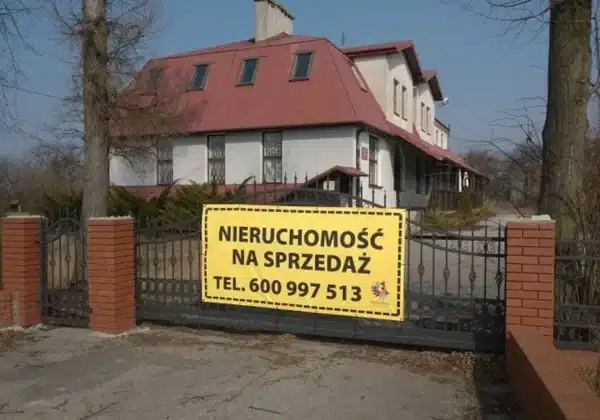 Na zdjęciu widać budynek mieszkalny z widocznym ogrodzeniem oraz dużym, wyraźnym banerem informującym o sprzedaży nieruchomości.