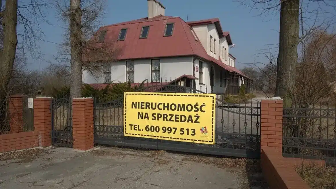 Na zdjęciu widać budynek mieszkalny z widocznym ogrodzeniem oraz dużym, wyraźnym banerem informującym o sprzedaży nieruchomości.