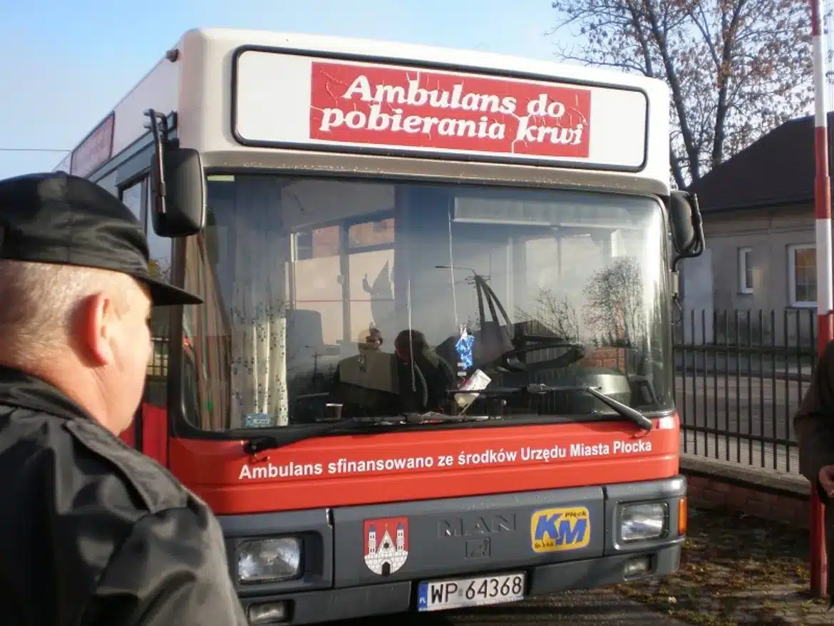 Na zdjęciu widoczny jest ambulans do pobierania krwi z napisem „Ambulans do pobierania krwi” oraz osoby zgromadzone wokół niego, w kontekście akcji pomocy chorej na białaczkę.