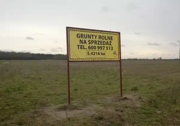 Na zdjęciu znajduje się duża, otwarta przestrzeń gruntów rolnych na sprzedaż z widocznym żółtym ogłoszeniem.