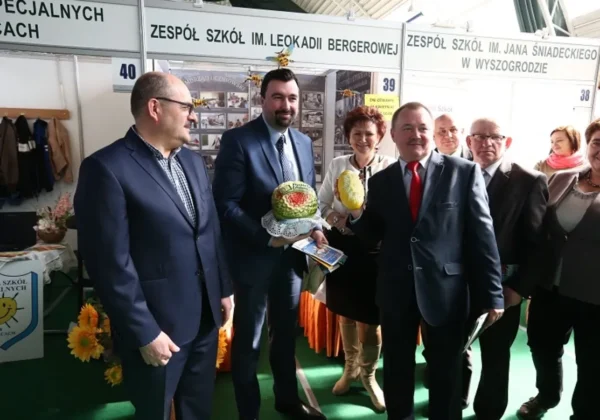 Grupa osób w garniturach na stoisku edukacyjnym podczas Płockich Targów Edukacyjnych, prezentujących dekoracyjne owoce i materiały promocyjne.