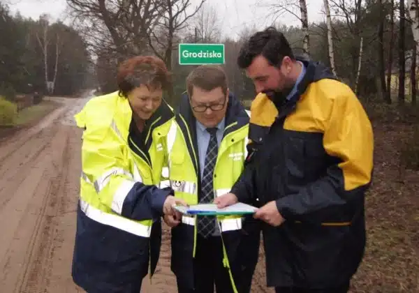 Na zdjęciu trójka osób w odzieży roboczej stoi na drodze, przeglądając mapę i omawiając kwestie dotyczące powiatowych dróg.