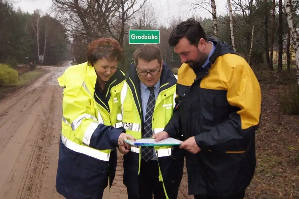Na zdjęciu trójka osób w odzieży roboczej stoi na drodze, przeglądając mapę i omawiając kwestie dotyczące powiatowych dróg.