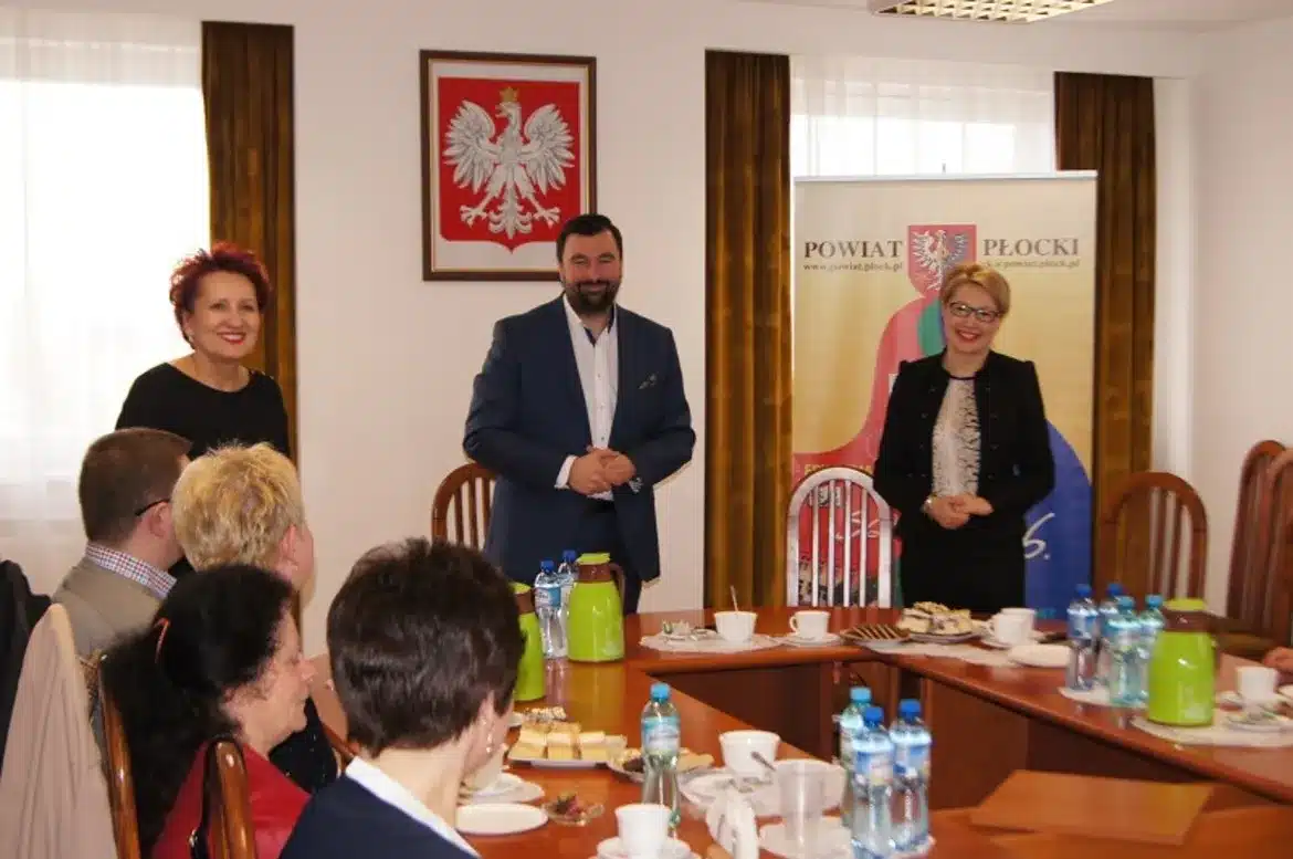 Spotkanie integrujące organizacje pozarządowe działające w sferze publicznej na terenie powiatu płockiego, z widocznymi uczestnikami przy stole oraz banerem powiatu w tle.