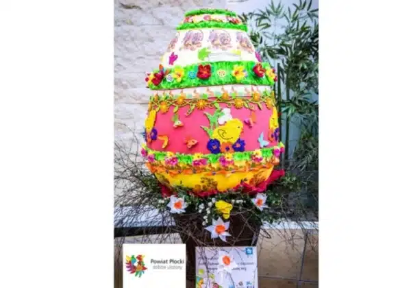 Na zdjęciu przedstawiona jest kolorowa, zdobiona pisanka umieszczona na stelażu w otoczeniu kwiatów, z okazji finału konkursu „Na najpiękniejszą pisankę” w płockim ZOO.