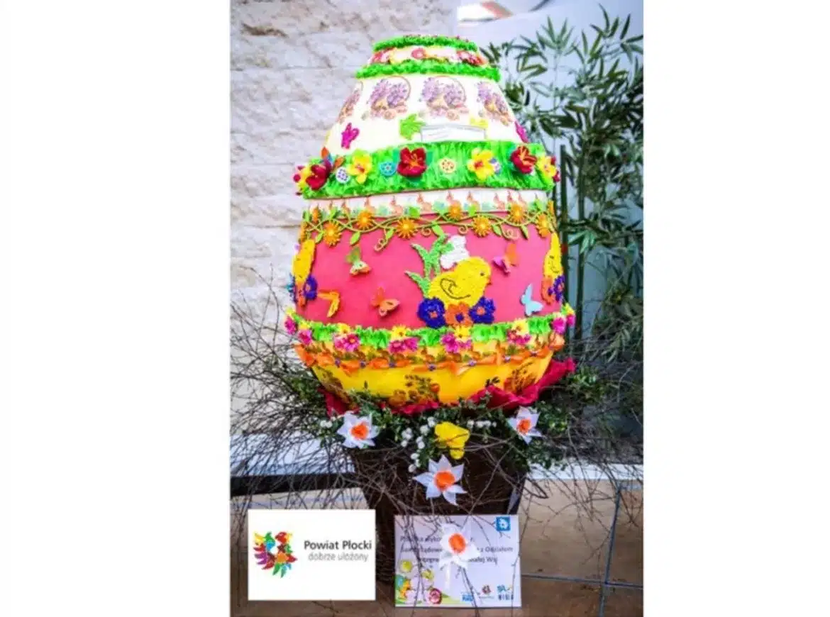 Na zdjęciu przedstawiona jest kolorowa, zdobiona pisanka umieszczona na stelażu w otoczeniu kwiatów, z okazji finału konkursu „Na najpiękniejszą pisankę” w płockim ZOO.