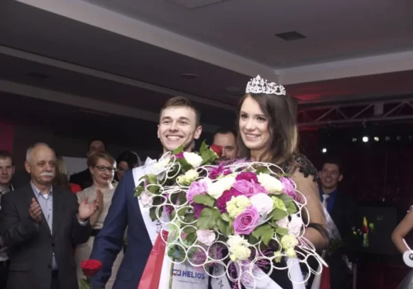 Na zdjęciu widać uśmiechniętą parę laureatów wyborów MISS i MISTERA PWSZ w Płocku, trzymających bukiety kwiatów, w otoczeniu zadowolonej publiczności.