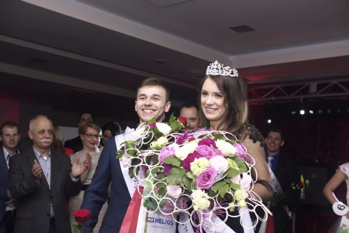 Na zdjęciu widać uśmiechniętą parę laureatów wyborów MISS i MISTERA PWSZ w Płocku, trzymających bukiety kwiatów, w otoczeniu zadowolonej publiczności.
