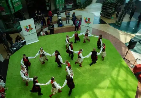 Dzieci tańczą w tradycyjnych strojach ludowych na zielonej podłodze podczas III Edycji Festiwalu Tańca i Zieleni w Galerii Mazovia, otoczone przez licznych widzów.