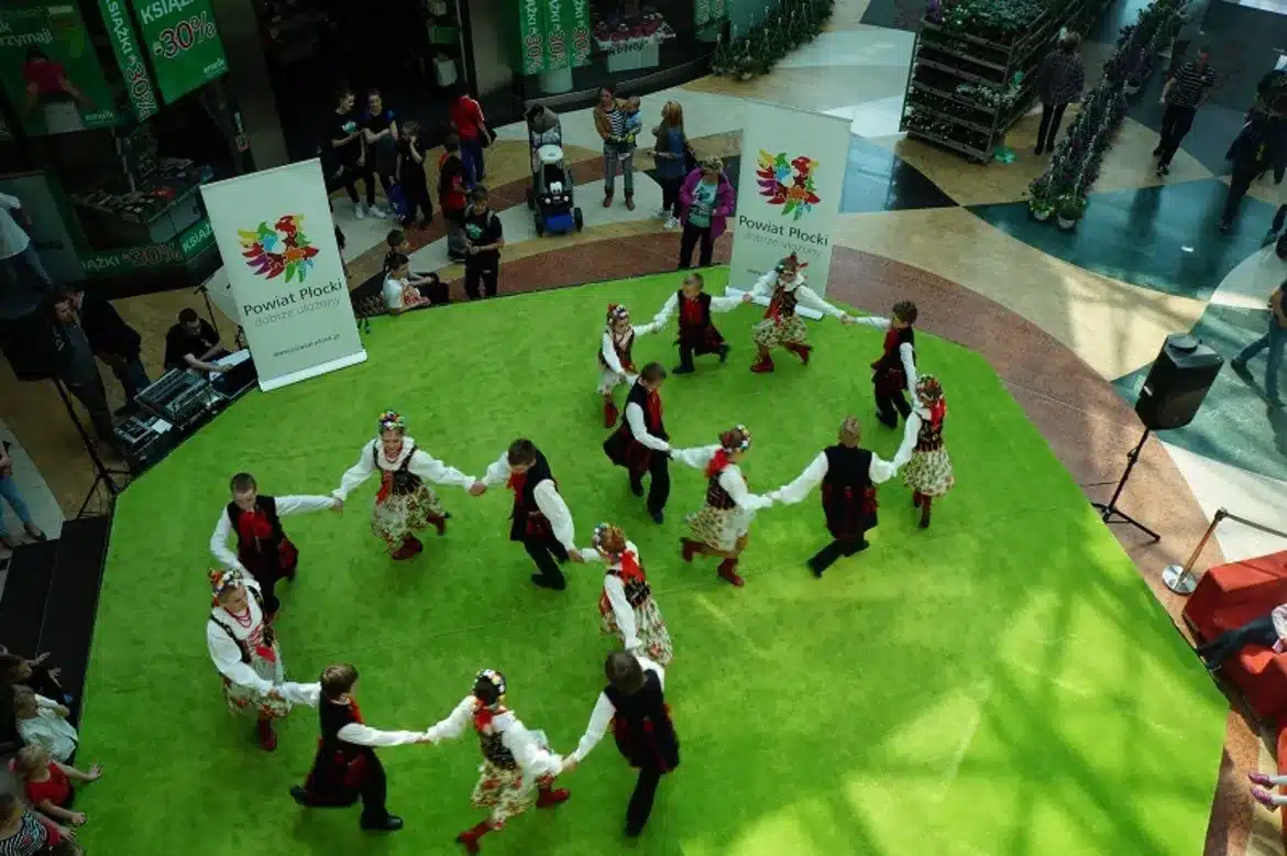 Dzieci tańczą w tradycyjnych strojach ludowych na zielonej podłodze podczas III Edycji Festiwalu Tańca i Zieleni w Galerii Mazovia, otoczone przez licznych widzów.