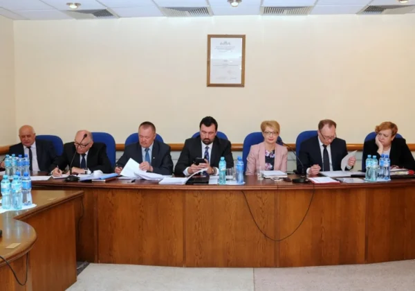 Siedmioosobowa grupa radnych i urzędników siedzi przy długim drewnianym stole z dokumentami, mikrofonami i butelkami wody podczas V Sesji Rady Powiatu w Płocku.