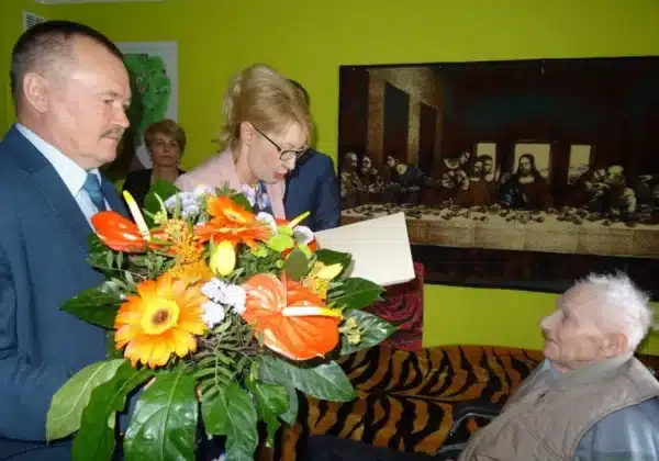Uroczystość z okazji setnych urodzin, podczas której jubilat otrzymuje kwiaty i gratulacje od gości.