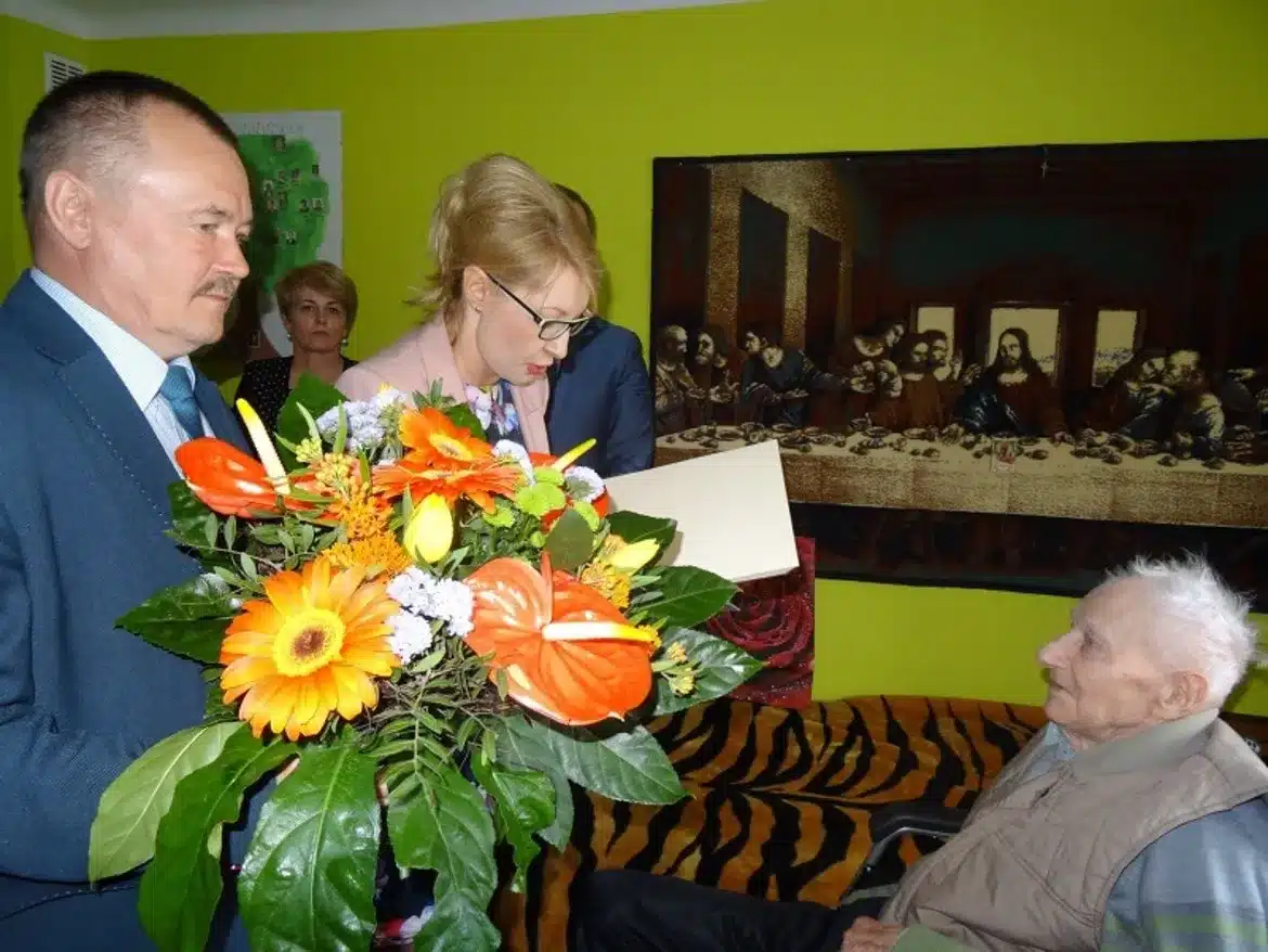 Uroczystość z okazji setnych urodzin, podczas której jubilat otrzymuje kwiaty i gratulacje od gości.