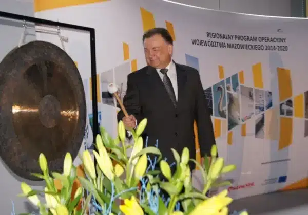 Na zdjęciu przedstawiono uśmiechniętego mężczyznę w garniturze, który trzyma pałeczkę i przygotowuje się do uderzenia w gong podczas wydarzenia związanego z Regionalnym Programem Operacyjnym Województwa Mazowieckiego na lata 2014-2020.