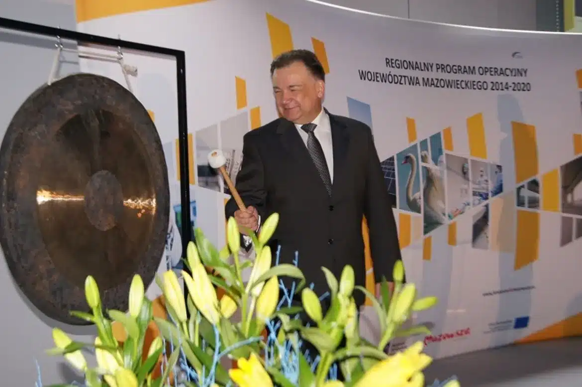 Na zdjęciu przedstawiono uśmiechniętego mężczyznę w garniturze, który trzyma pałeczkę i przygotowuje się do uderzenia w gong podczas wydarzenia związanego z Regionalnym Programem Operacyjnym Województwa Mazowieckiego na lata 2014-2020.