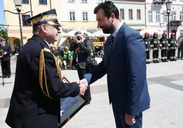 Uroczystość z okazji Dnia Strażaka oraz 140-lecia Straży Ogniowej w Płocku, z uściskiem dłoni na tle strażaków w mundurach.