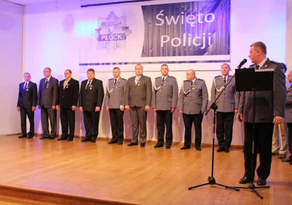 Grupa policjantów w mundurach i odznaczeniach stoi na scenie podczas Święta Policji w Płocku, a po prawej stronie stoi oficer przy mikrofonie.