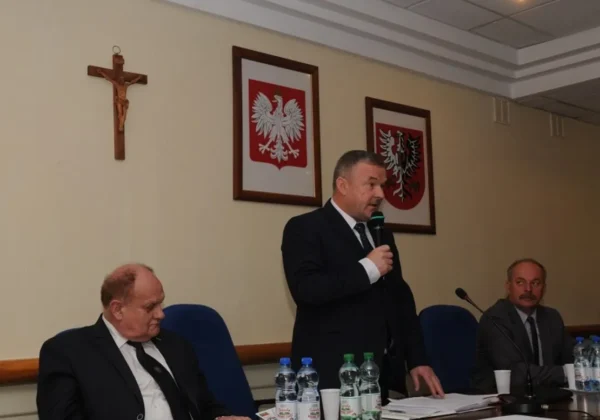 Mężczyzna w garniturze przemawia do mikrofonu podczas VIII Sesji Rady Powiatu Płockiego, stojąc przy stole obok dwóch innych mężczyzn; w tle widoczne krzyż i dwa oprawione herby państwowe.
