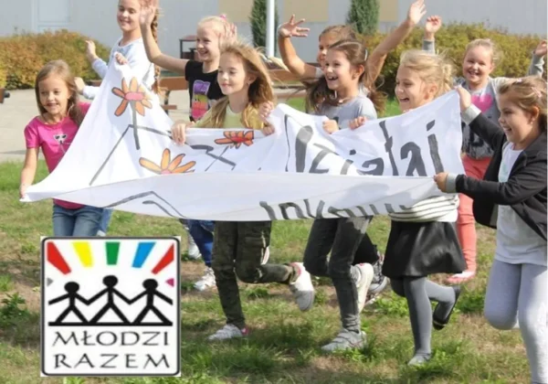 Grupa dzieci biegnie na zewnątrz, trzymając duży biały baner z rysunkami kwiatów; w dolnym lewym rogu widoczne jest logo Młodzi Razem Funduszu Lokalnego Ziemi Płockiej.
