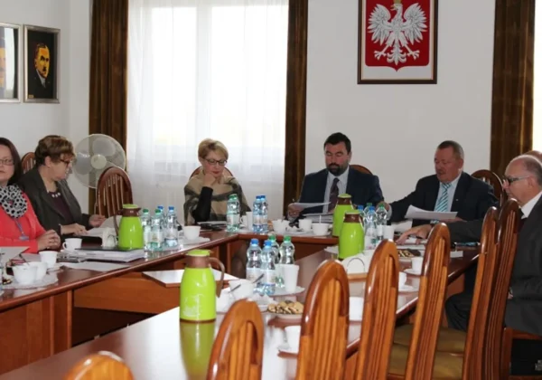 Członkowie Zarządu Powiatu w Płocku siedzą przy długim stole konferencyjnym podczas posiedzenia, omawiają dokumenty, a na stole są napoje i przekąski, w tle widnieje godło Polski.