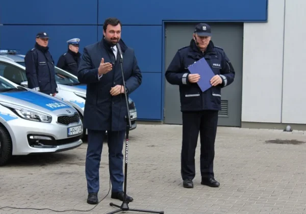 Na zdjęciu przedstawiono mężczyznę prowadzącego przemówienie przy mikrofonie, obok niego stoi policjant z dokumentem, a w tle widać radiowozy policyjne.