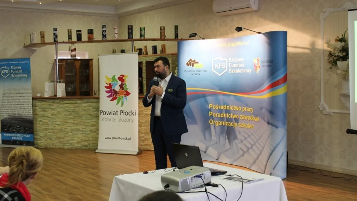 Na zdjęciu przedstawiono mężczyznę przemawiającego podczas prezentacji dotyczącej Krajowego Funduszu Szkoleniowego, z logo funduszu oraz Powiatu Płockiego w tle.