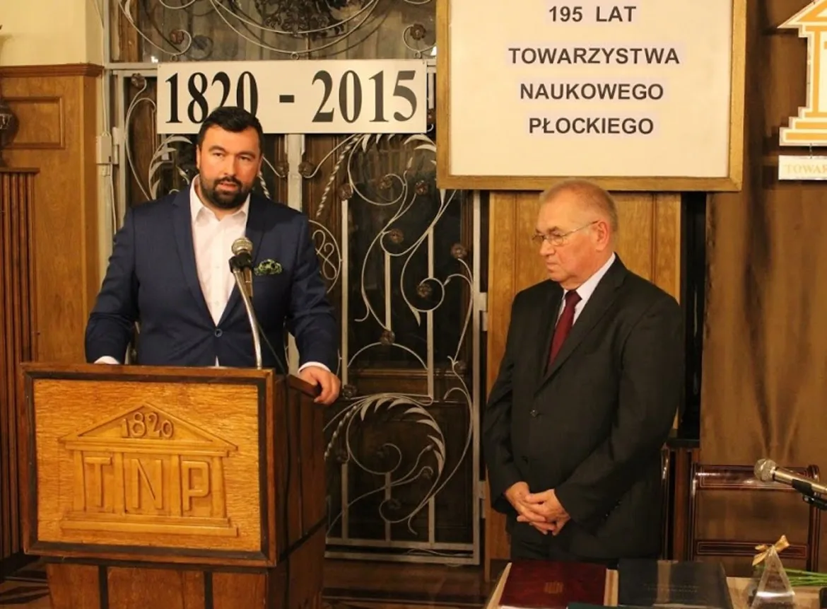 Zdjęcie przedstawia dwóch mężczyzn podczas uroczystości związanej z 195-leciem Towarzystwa Naukowego Płockiego, z jednym z nich mówiącym do mikrofonu.