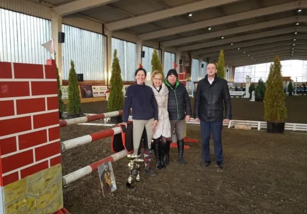 Cztery osoby stoją przy przeszkodzie na halowych zawodach towarzyskich w Stadzie Ogierów w Łącku, trzymając puchar i uśmiechając się do kamery.