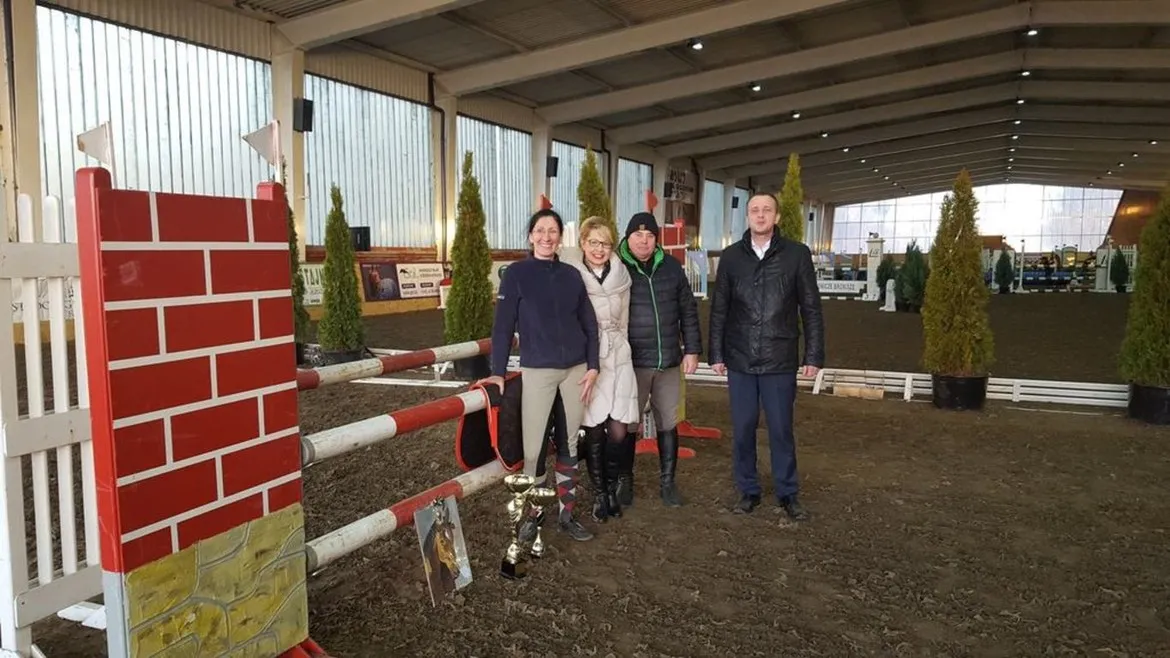 Cztery osoby stoją przy przeszkodzie na halowych zawodach towarzyskich w Stadzie Ogierów w Łącku, trzymając puchar i uśmiechając się do kamery.