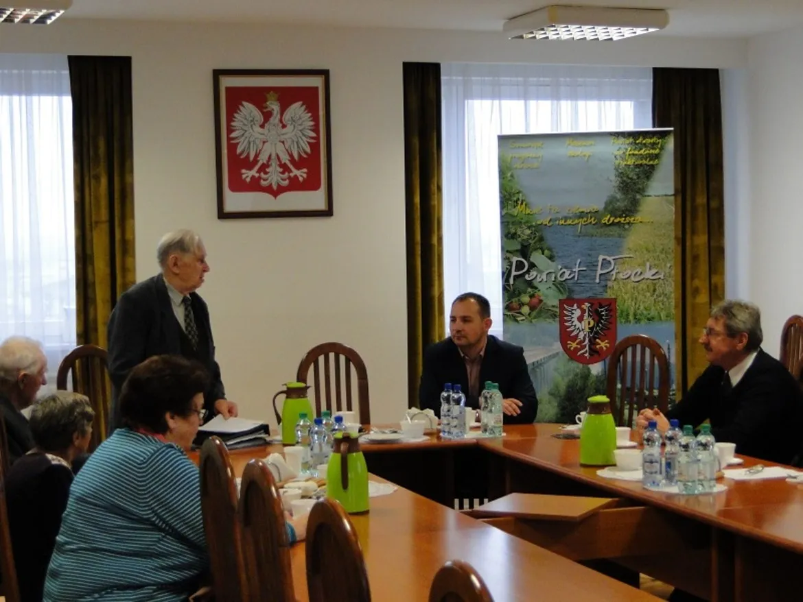 Posiedzenie Zarządu Koła Powiatowego Kombatantów i Osób Represjonowanych, w którym uczestniczą starsi mężczyźni i kobiety, dyskutując w sali z elementami dekoracyjnymi na tle.