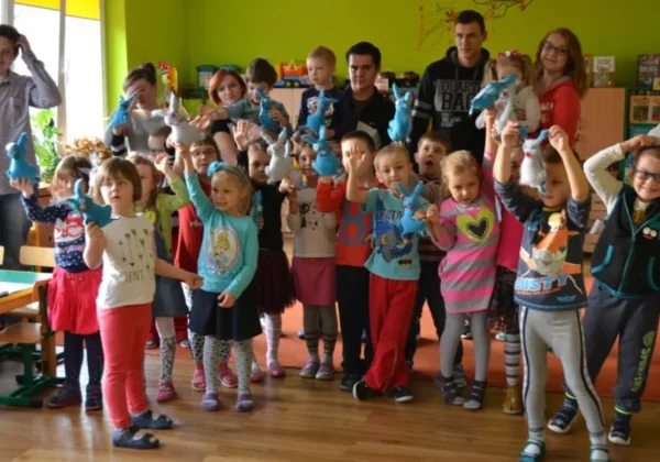 Dzieci i młodzież w kolorowej klasie trzymają w górze niebieskie maskotki oraz uśmiechają się w ramach projektu Szkolnego Klubu Wolontariusza „Jestem dla innych”.