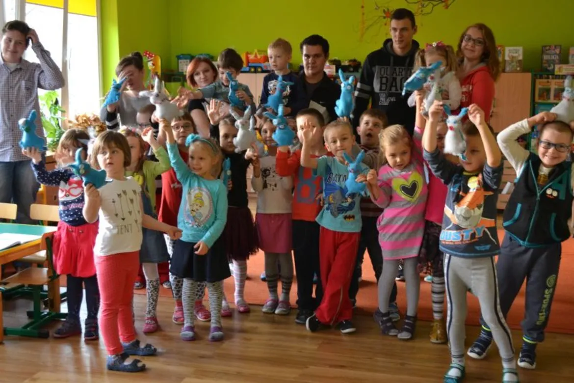 Dzieci i młodzież w kolorowej klasie trzymają w górze niebieskie maskotki oraz uśmiechają się w ramach projektu Szkolnego Klubu Wolontariusza „Jestem dla innych”.