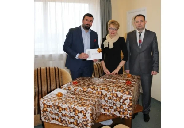 Trzy osoby stoją przy stoliku z ozdobnie zapakowanymi paczkami w ramach akcji Szlachetna Paczka, prezentując także kartkę z napisem.