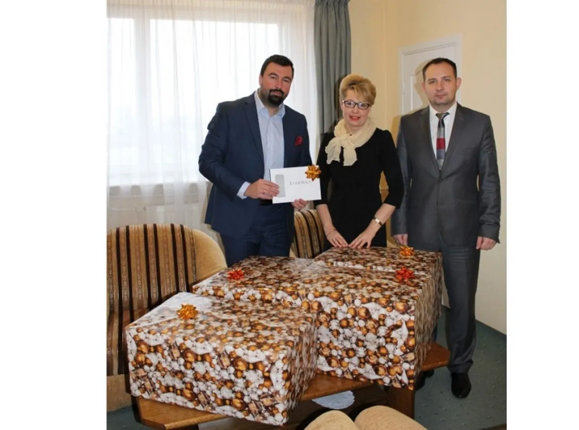 Trzy osoby stoją przy stoliku z ozdobnie zapakowanymi paczkami w ramach akcji Szlachetna Paczka, prezentując także kartkę z napisem.