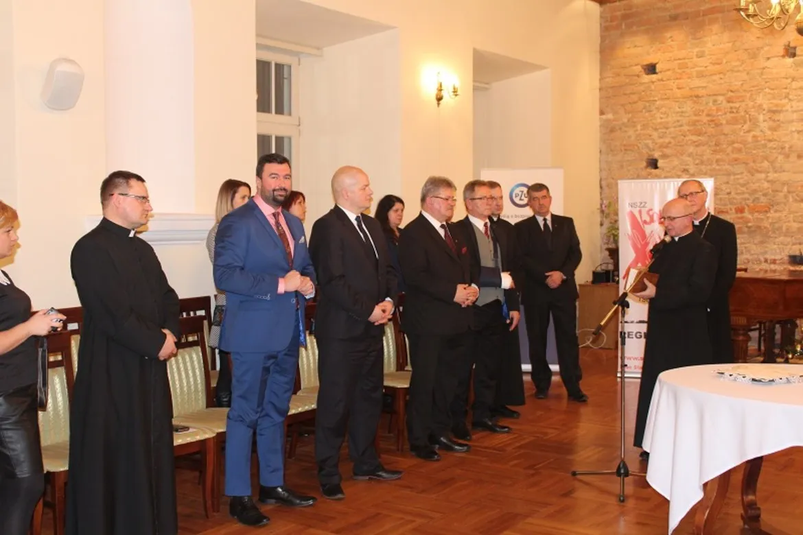 Spotkanie opłatkowe Płockiej Solidarności, z uczestnikami stojącymi w eleganckiej sali, w otoczeniu dekoracji świątecznych.