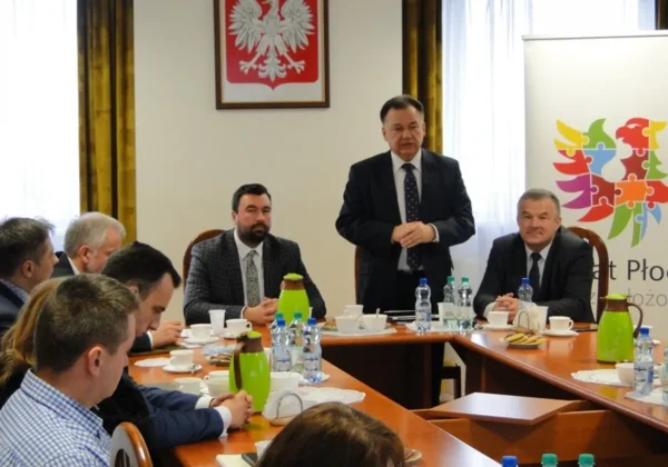 Na zdjęciu przedstawiona jest konferencja w Powiecie Płockim, gdzie mężczyzna w garniturze wygłasza przemówienie do zgromadzonych uczestników przy okrągłym stole.