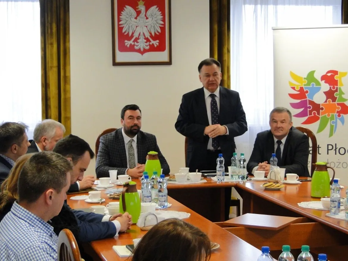 Na zdjęciu przedstawiona jest konferencja w Powiecie Płockim, gdzie mężczyzna w garniturze wygłasza przemówienie do zgromadzonych uczestników przy okrągłym stole.