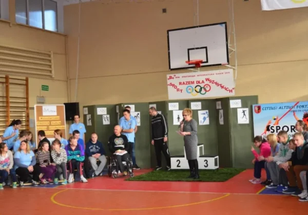 Uczestnicy wydarzenia zorganizowanego w ramach Rządowego Programu „BEZPIECZNA +” gromadzą się na sali sportowej, gdzie jedna z prowadzących przemawia do zebranych, a w tle widoczne są medale i symbole olimpijskie.