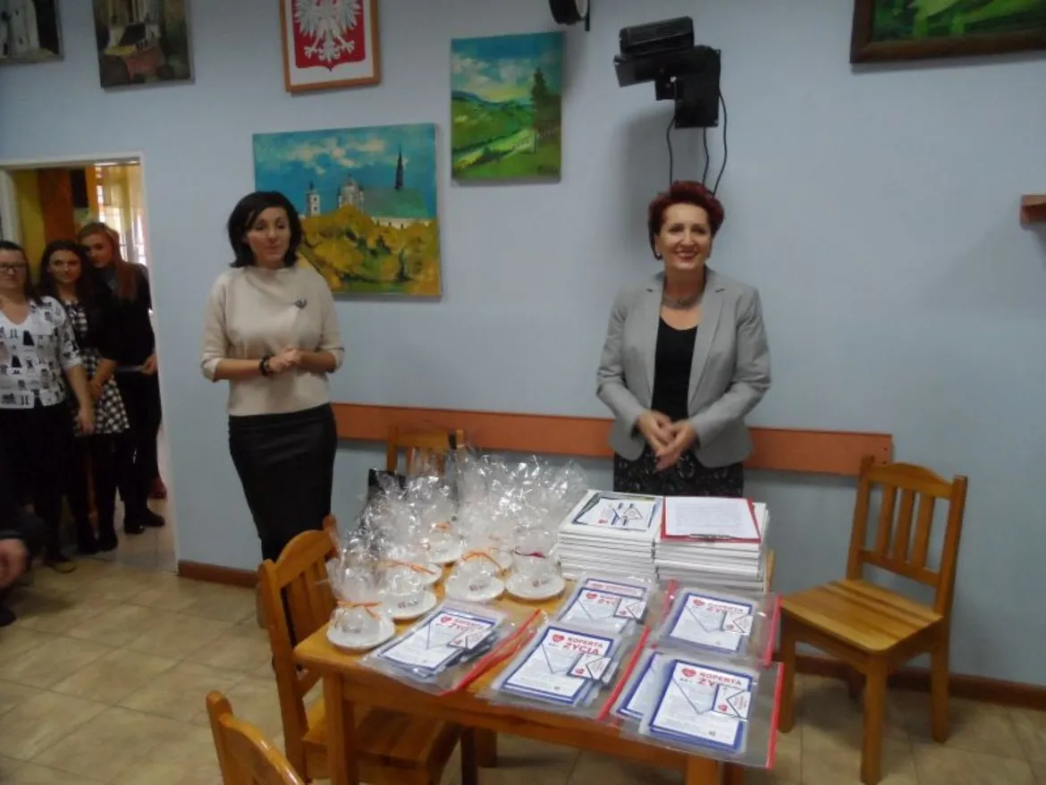 W spotkaniu na temat „Koperty Życia” uczestniczą dwie kobiety, podczas gdy zebrani słuchają o inicjatywie mającej na celu wsparcie osób potrzebujących.