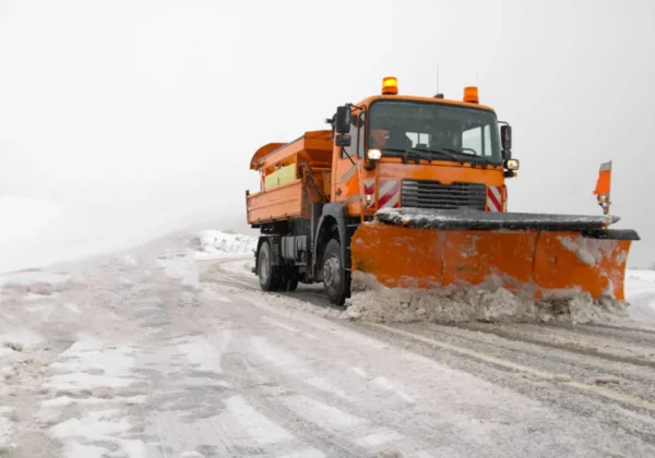 Na zdjęciu widoczny jest pomarańczowy samochód ciężarowy z pługiem śnieżnym, który odśnieża zaśnieżoną drogę w zimowej scenerii powiatu płockiego.