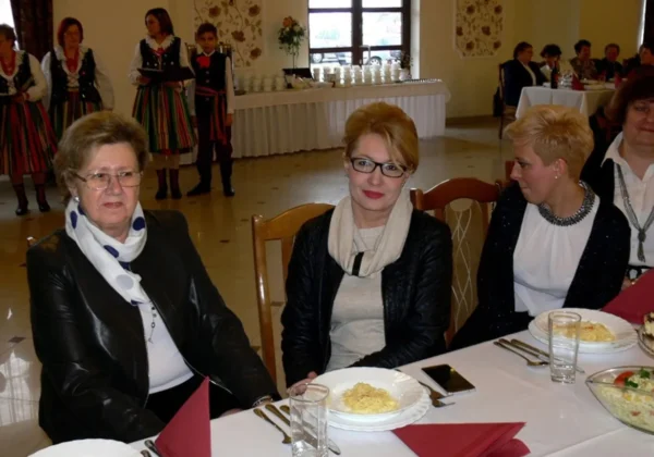 Na zdjęciu przedstawiono grupę uczestników XVII spotkania pokoleń w gminie Słubice, którzy siedzą przy stole w towarzyskim klimacie, a w tle widoczni są występujący w strojach ludowych.