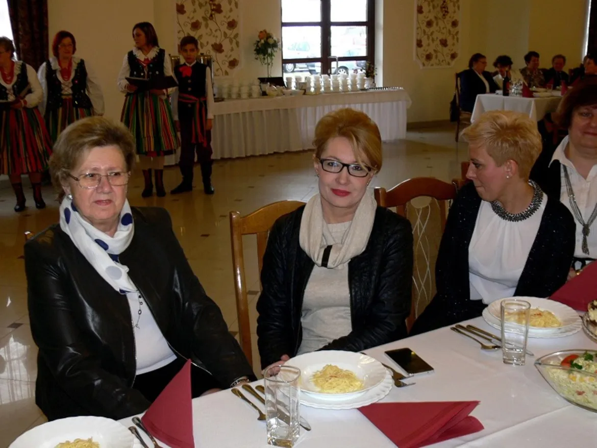 Na zdjęciu przedstawiono grupę uczestników XVII spotkania pokoleń w gminie Słubice, którzy siedzą przy stole w towarzyskim klimacie, a w tle widoczni są występujący w strojach ludowych.