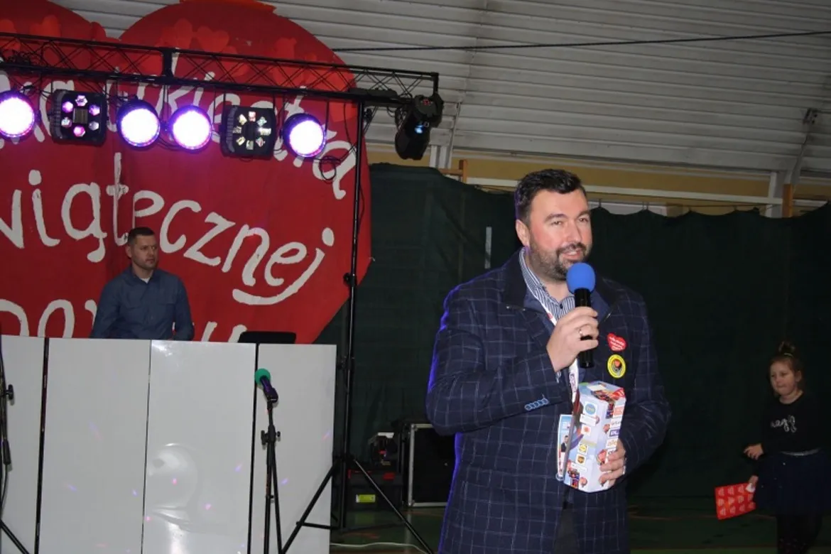 Na zdjęciu przedstawiono mężczyznę z mikrofonem, który przemawia podczas wydarzenia organizowanego przez Wielką Orkiestrę Świątecznej Pomocy, w tle widać scenę z oświetleniem oraz dekorację w kształcie serca.